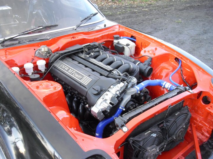 Project Datsun 280ZX - E36 M3 Evo Engine Swap - Page 6 - Readers' Cars - PistonHeads - The image shows a car with its engine compartment open, revealing a large array of mechanical components. This appears to be a classic or vintage car, indicated by the orange painted parts of the engine. The vehicle is parked on a street or driveway with a paved surface visible. The engine itself is quite large and includes various tubes, air filters, and other automotive hardware. The focus of the image is the intricate details of the engine, offering a glimpse into the mechanical complexity of vintage automobiles.