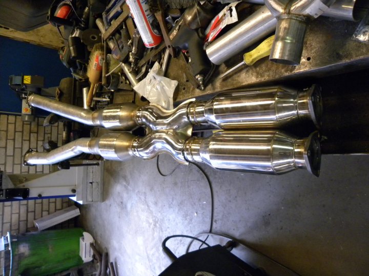 Pistonheads - The image shows a well-equipped metal shop filled with various tools and equipment. There's a large workspace dominated by a multitude of extruded metal tubing with specific designs, likely indicating some kind of engineering or metalworking project. There are large, dirty pipe sections that suggest heavy-duty work, and power tools like a sander and hammers are scattered across the room, ready for use. The background is mostly obscured, but a brick wall and some human figures can be seen, possibly indicating the presence of a workforce in the workshop.