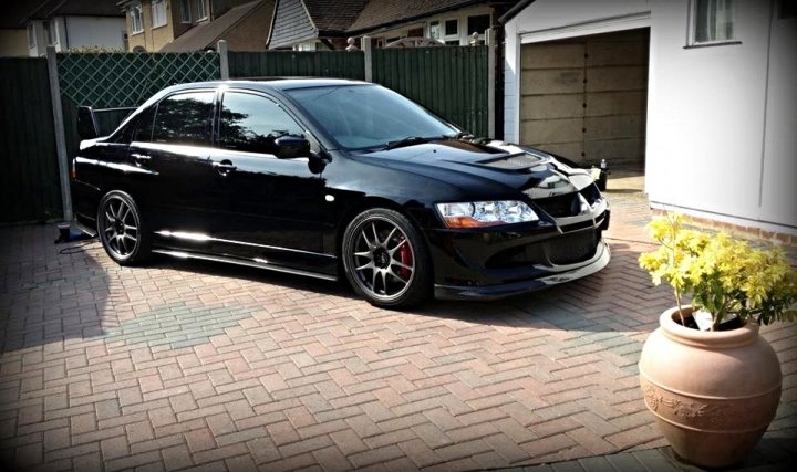 Post your Evo pics - Page 1 - Jap Chat - PistonHeads - The image shows a sleek black car parked in a driveway with a large potted plant to the right. The car has a specialized front bumper that suggests it is a modified or racetrack car. The driveway appears to be made of red brick and there is a wooden gate behind the car. In the background, there's a neatly trimmed yard and part of a white house can be seen. The setting seems to be a residential area with a focus on the unique features of the car.