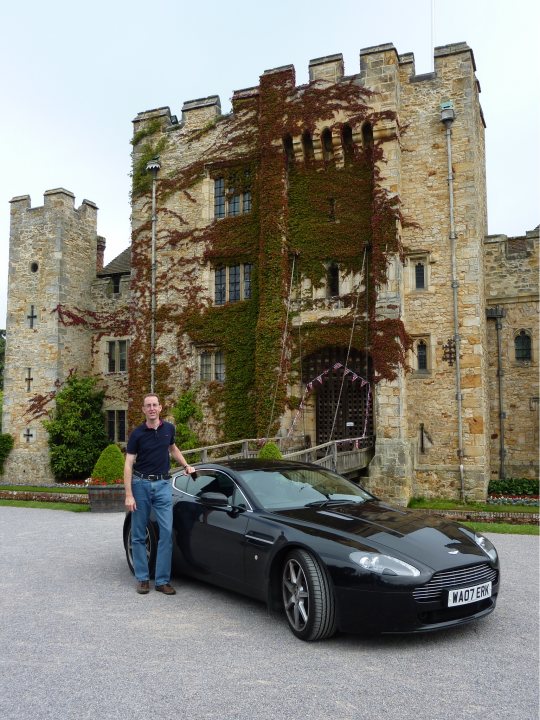 Trip of a Lifetime (picture heavy) - Page 1 - Aston Martin - PistonHeads - The image depicts a man standing next to a sleek black sports car, possibly an Aston Martin, in front of a grand, historic mansion or castle. The car is black and shiny, exuding a sense of luxury and power. The man is posing, and there is a water fountain in the foreground. The mansion boasts a towering and intricate architectural structure, likely constructed from stone, and the front is adorned with lush greenery, including a large ivy plant climbing the wall. The setting is serene and majestic, suggesting a peaceful or meticulously maintained estate.