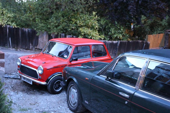 It followed me home, can we keep it? - Page 4 - Rolls Royce & Bentley - PistonHeads - The image shows a gravel driveway with two distinctly different cars. On the left, there's a vibrant red Mini Cooper, positioned near the fence. To the right, there's a classic black Jaguar with a silver top, partially obscured by the red car to its left. The scene takes place in a residential setting, with trees and bushes visible in the background. There are a few elements of wear and tear on the cars, suggesting that they've been used for a considerable amount of time.