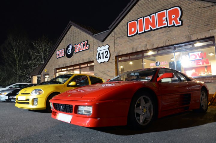 Pistonheads Essex March - The image depicts a scene on a dark night, outside a red brick building named "Diner". A vibrant red Ferrari sports car from the 1980s takes center stage, parked in front of the diner, exuding a sense of luxury and speed. A yellow SmART car is situated to the left of the Ferrari, adding a pop of contrasting color. The building itself has a red awning, enhancing the overall red theme, and is illuminated by white and red lights, casting a warm glow on the vehicles and the exterior. The number '412' is prominently displayed on the facade of the diner, possibly indicating its location or route. The image captures the essence of nightlife, with the vintage design of the diner and the modernity of the sports cars intertwining.