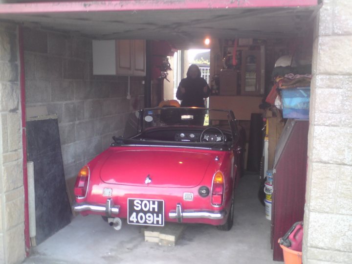 Pistonheads - The image shows a vintage red car parked inside a garage with a sloped roof. The interior light of the car appears to be on, illuminating the front interior. A person is partially visible on the right side of the image, standing and facing the car. The garage setting features a concrete floor and a bulkhead at the back of the garage. Various items such as a cardboard box and a yellow ABSOS container can be seen in the background.