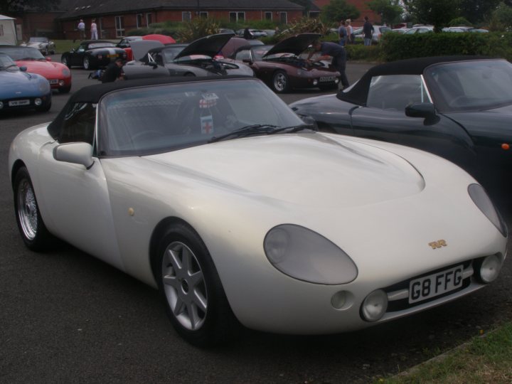 Pistonheads Griff Growl - The image features a vintage white sports car prominently from the side, parked in a lot with several other cars. The sports car is characterized by its smooth curves, prominent side scoops, and large headlights. It's covered by a car cover. The rest of the cars in the background appear to be modern or of different styles. The setting seems to be an urban parking lot, given the presence of buildings and a paved surface. There are people in the background, likely pedestrians, which adds a sense of scale to the image.