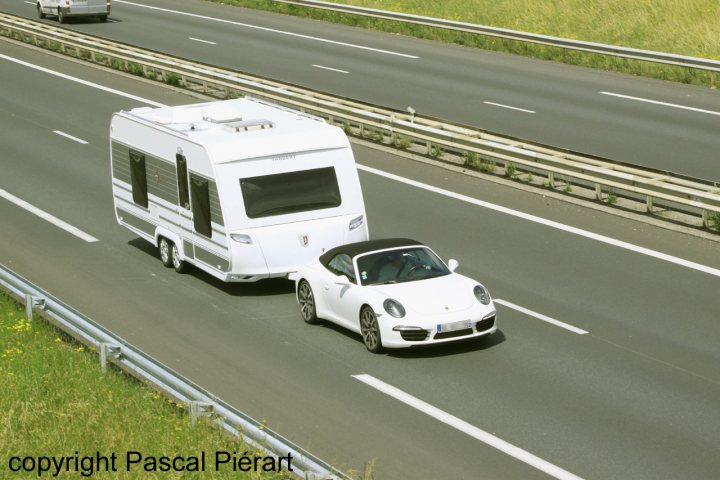 Pistonheads - The image depicts a two-lane highway with traffic. On the right lane, there is a white car with its roof open, driving towards the camera. In the left lane, following slightly behind, there is a silver motorhome or RV. There's also another smaller car in front of the RV. The vehicles are traveling on a clear day, and the highway appears to be free of other cars and trucks, allowing these two vehicles to drive unobstructed. The background shows an open landscape with no immediate buildings or structures visible, giving the impression of being in a rural or less populated area.