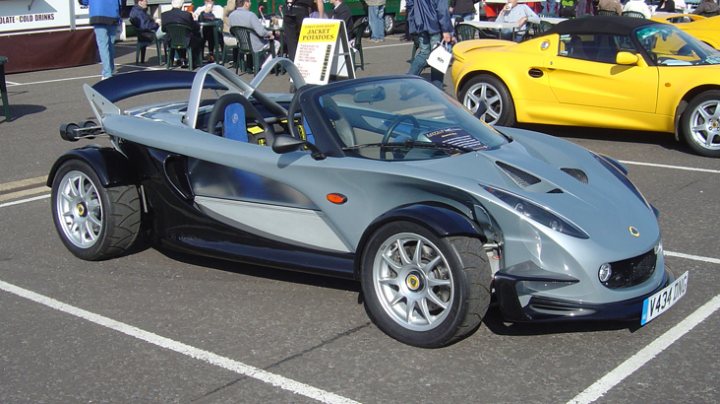 Caterham Readying Pistonheads - The image captures a scene of a futuristic-looking sports car parked in a lot amidst other conventionally designed cars. The sports car stands out due to its sleek, silver-gray exterior and aerodynamic design. The lot appears to be a public parking area with several spots occupied by other cars. The surrounding environment gives the impression of an outdoor exhibition, possibly a car show, with various blue and white signs visible in the background.