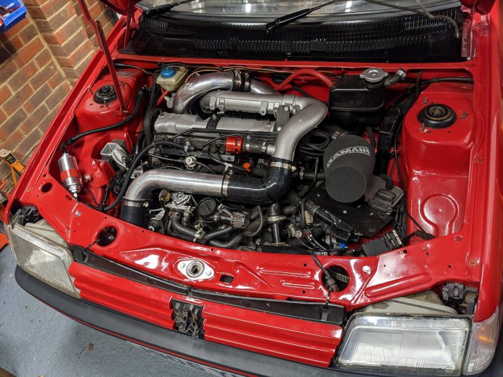 205 GTi Lemon!  - Page 1 - Readers' Cars - PistonHeads UK - The image showcases an open red hatchback car in a garage, with the hood up revealing its engine. Various mechanical components are visible under the hood, including what appears to be a turbocharger and intake system. A pipe, possibly part of the car's exhaust system, is prominently displayed. The car has a damaged front bumper, indicating it may have been involved in an accident or has experienced some form of collision damage.