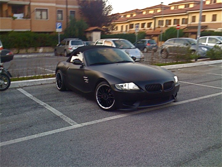 Pistonheads Wrap Zmc Vinyl Carbon - The image showcases a black BMW parked in a parking lot. It has a sleek and modern design, characterized by a long hood, short rear deck, and a distinctive BMW kidney grille. The car is positioned at an angle and faces the left side of the image. The car is parked alongside white parking lines, opposite another row of parked cars. The setting appears to be an urban environment with buildings visible in the background.