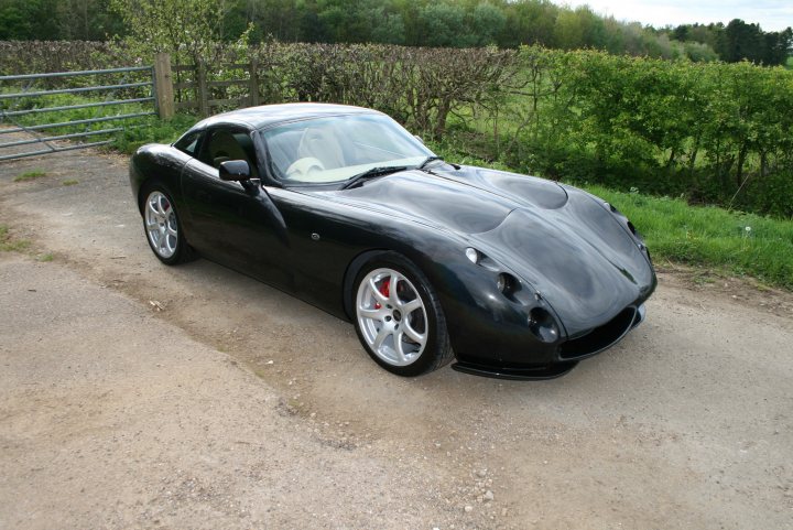 Pistonheads Tvr Tuscan - The image showcases a classic black sports car, parked on a gravel road. The car's sleek design and shiny black finish give it an aggressive yet elegant look. It is positioned in front of a lush green fence, which contrasts nicely with the car's color. The surrounding environment includes trees and a grassy area, suggesting a peaceful, rural setting. Features such as the distinctive headlamps, air vents, and the overall shape of the car indicate it is a specific model, possibly an early 2000s vehicle.