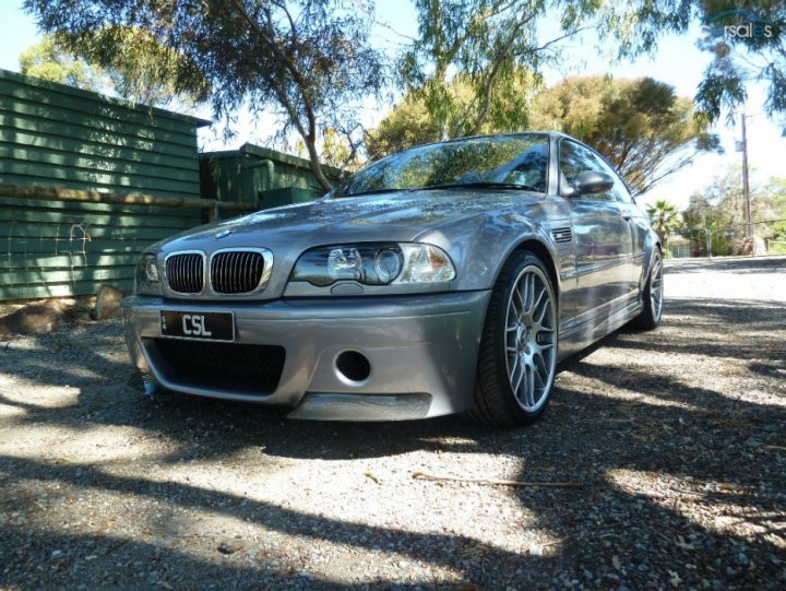 A white car parked in a parking lot - Pistonheads - This image shows a silver-colored compact car parked on what appears to be a gravel driveway. The car is positioned at an angle to the camera, revealing parts of its front and side. It has a prominent front grille with a model emblem, and the rims on alloy wheels are highly detailed. The car is not evidently in motion. The vehicle's position at the top of the image suggests it may be parked on an elevated area. There are trees and a green fence in the background, and the lighting conditions indicate it's a clear day.