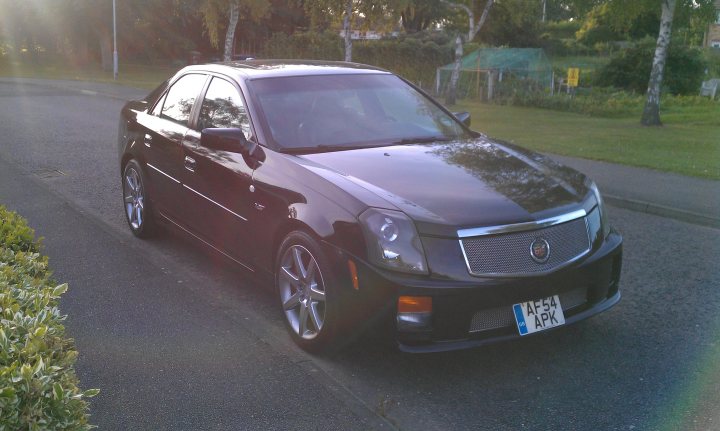 2005 CTS-V - Page 1 - Yank Motors - PistonHeads - The image showcases a black sedan parked on the side of a road during daylight. The car is positioned at an angle from the foreground, allowing a clear view of its front end and part of its side profile. It has a polished exterior reflecting the sunlight, and the photograph has captured a vibrant lens flare on the right side, adding to the overall aesthetic. The vehicle also displays a European-style license plate, which, along with the distinctive design, suggests that the car might be from a European manufacturer.