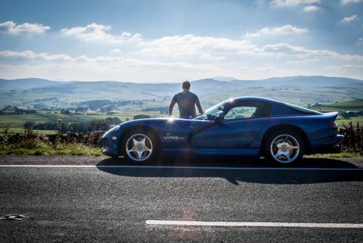 Evo Triangle (pics and vid) - Page 1 - Vipers - PistonHeads - In the image, a man stands on the side of a road, overlooking a picturesque landscape. He is positioned in front of a sleek, blue sports car, which is parked along the edge of the pavement. The man appears contemplative, with his gaze directed out over the lush green hills and valleys that spread out beneath him. The sky is clear, suggesting a bright, sunny day. The setting suggests a moment of quiet appreciation of nature and a love for fast automobiles. The environment is peaceful and calm.