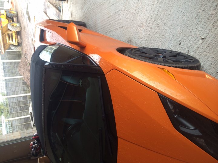 More pics of my LP560 - Page 1 - Gallardo/Huracan - PistonHeads - The image shows an orange car positioned in a parking lot. The car's design includes black accents and its hood is propped open, revealing a partially visible engine. It seems to be raining, as the ground is wet and there are drops of water on the car's surfaces. In the background, there are structures with bars on them, indicating a location likely to be used for storage or industrial purposes. There is also a piece of machinery, possibly construction equipment, visible near the car, suggesting an industrial or commercial setting.