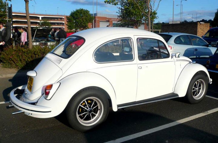 Pistonheads - The image features a white VW Beetle parked in a parking lot with other cars in the background. The car's condition suggests it is well maintained, with a noticeable chip on the side of its body. The setting is outdoors, likely in an urban environment, as there are buildings and cars in the vicinity. There are people near the parked cars, suggesting a bustling everyday scene.