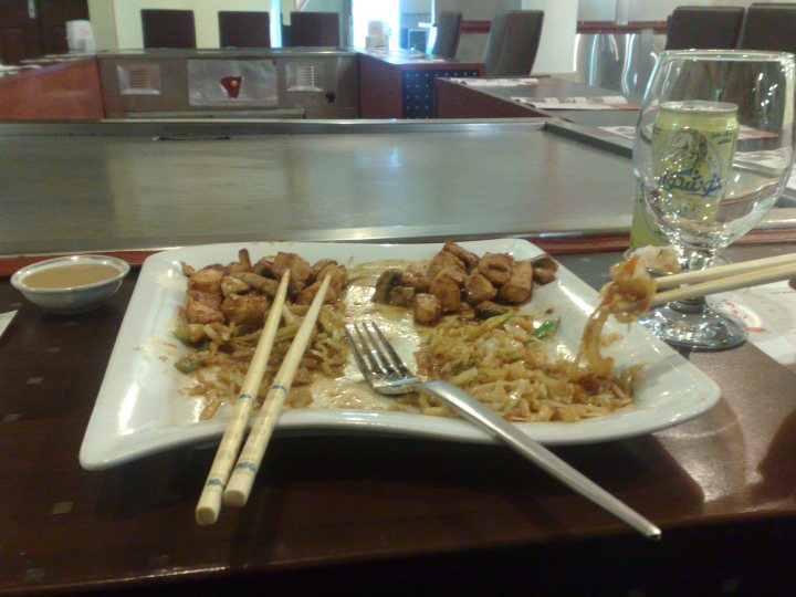 The image presents a vibrant dining scene featuring a variety of Asian cuisine on a white rectangular plate. The main dish consists of a combination of stir-fried noodles, which appear to be szechuan noodles, and a mix of semi-sliced, possibly bird, meat. The plate is further garnished with a pair of white chopsticks resting on it. In the background, there is a dining table with chairs, and other food items can be seen, including dumplings, which are elevated above the central dish, making the noodles the focal point of the image. The setting suggests a casual dining environment.