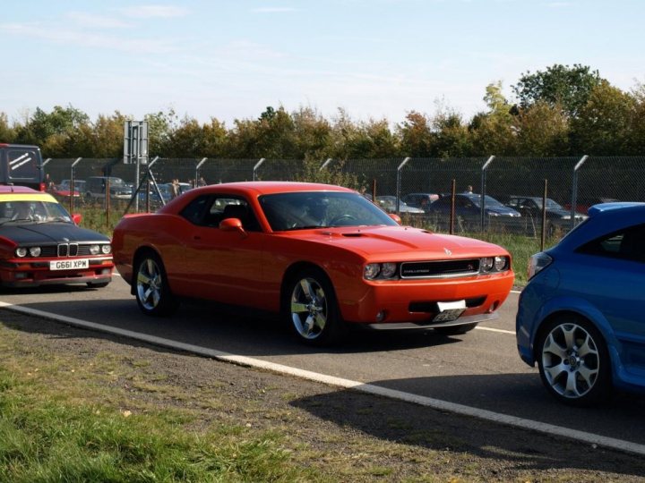 Dodge Revealed Charger Pistonheads - This image captures a scene on a roadway. There are two cars in the foreground; a vibrant red sports car is parked on the right side of the road, while a bright blue car is passing by on the left. The road stretches out in the background with a fence on one side. There are also some other vehicles and a person in the distance, adding depth to the scene.