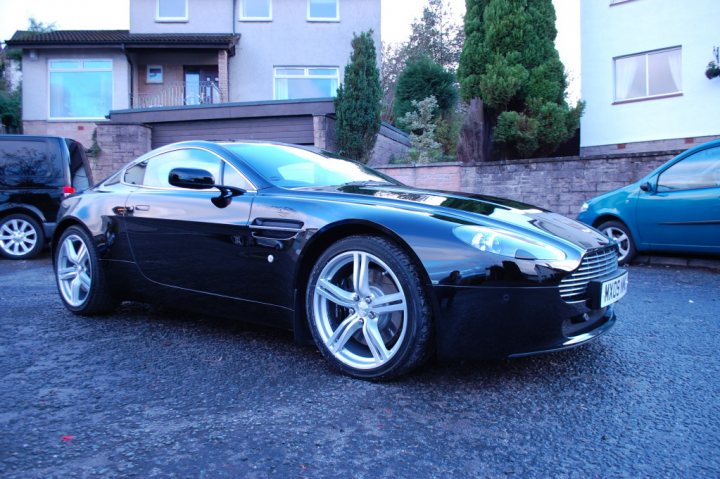 Pistonheads - The image features a black Aston Martin car parked on an asphalt surface. The car is positioned at a slight angle to the camera, highlighting its sleek design and shiny paintwork. In the background, there are two other cars parked to the left and a house with modern architecture is partially visible behind the Aston Martin. The sky is clear and the scene is taken during the day under blue, bright skies.