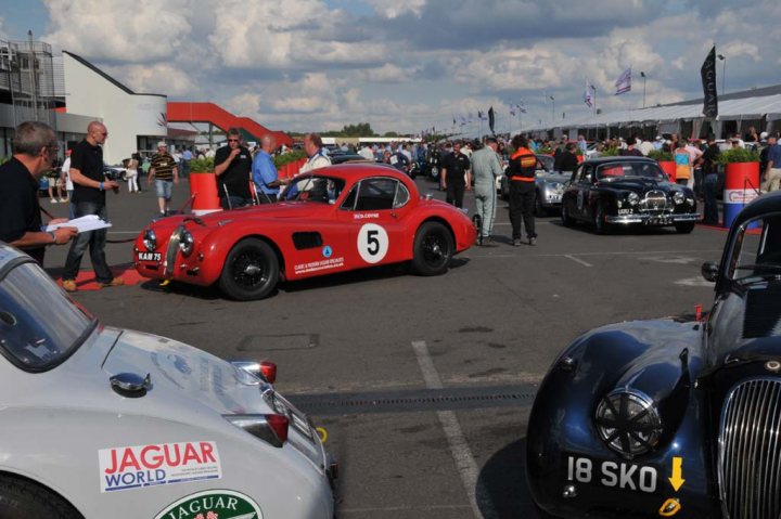 RW774  and the XKs at historic Silverstone - Page 1 - Classic Cars and Yesterday's Heroes - PistonHeads - The image shows a bustling scene at a classic car show. A vintage Jaguar and a sleek, vintage Jaguar's replacement are prominently featured in the foreground. These cars and several other classic cars are parked on either side of a large parking lot. Numerous people can be seen walking among the cars, inspecting them closely, and engaging in conversations. In the background, there are various structures that likely house the show-related activities and possiblyevent stalls. The sky is overcast with clouds, suggesting an outdoor setting and possibly indicating cool weather conditions.