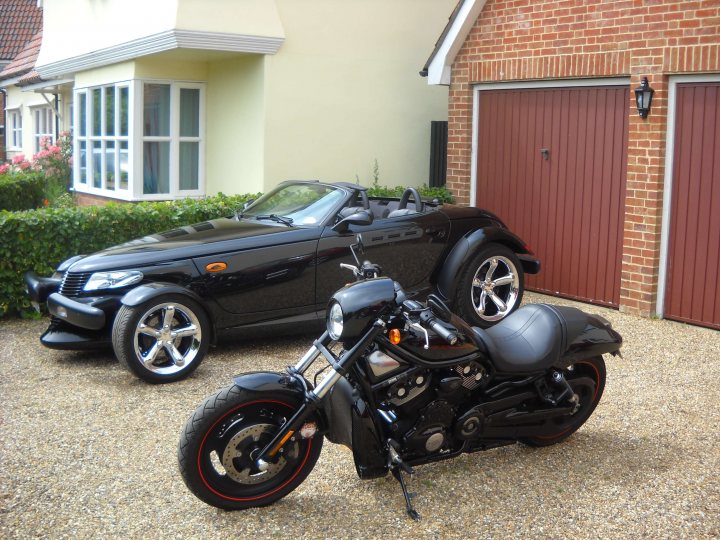 A picture a day....biker banter (Vol 2) - Page 358 - Biker Banter - PistonHeads - The image showcases a garage with two motorcycles parked outside. To the left is a black sports car with a convertible roof, and to the right is a black motorcycle with large rear fenders. The motorcycle has orange and red accents, predominantly in its tires. The background reveals a red brick house with a white trim above a row of bushes. The setting appears to be a quiet, suburban area with a paved driveway. The overall scene gives a sense of luxury and outdoor hobbies.