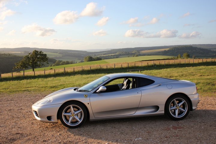 360 thread - Page 1 - Ferrari V8 - PistonHeads - The image showcases a silver Ferrari parked on a gravel road in a rural setting. The car is resting under a sunny sky, basking in the soft light. The surrounding landscape features a gentle hill, a fence, and lush greenery. In the distance, beyond the fence, lies a verdant valley that stretches towards the horizon. The Ferrari, with its sleek design and shiny silver color, stands out against the natural backdrop, creating a striking contrast between luxury and nature.