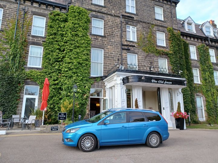 The image shows a scene with a car parked in front of a building. The building is two stories high and has several windows, some of which are covered with ivy. A red flag hangs from the entrance of the building. In front of the building, there's a small area with some plants and a bench. On the ground floor of the building, there is a sign that reads "The White Hart," suggesting it could be a hotel or restaurant. The car parked in front appears to be a blue compact vehicle. The street on which this scene takes place has a brick sidewalk and a lamp post, typical elements of an urban setting.