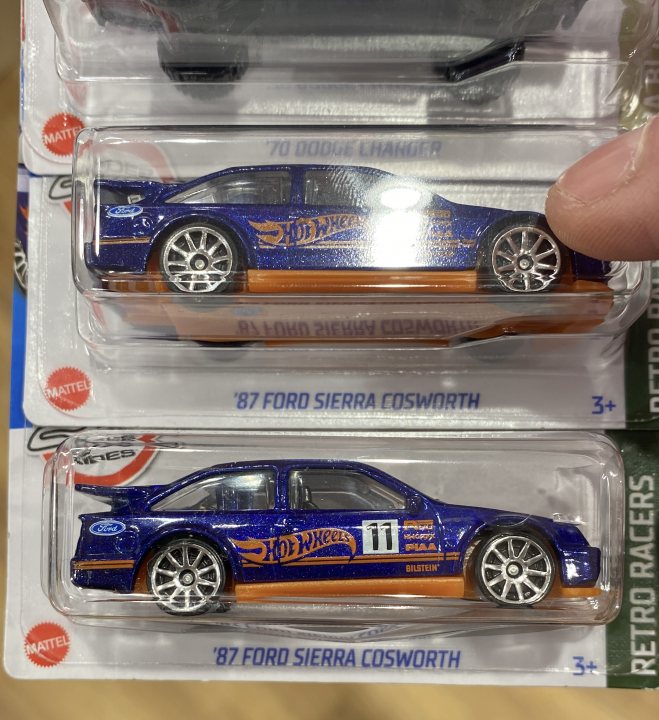 Hot Wheels 1:64 - Page 4 - Scale Models - PistonHeads UK - The image shows a collection of three Hot Wheels cars displayed on a flat surface. Each car has its own packaging, with the top car labeled "70 Ford Sierra Cosworth" and the other two labeled "81 Ford Sierra RS500." There's a hand visible in the frame, likely holding or inspecting one of the cars. The Hot Wheels branding is clearly visible on the packaging. The overall setting appears to be indoors, as suggested by the plain wall in the background. The focus of the image is on the toy cars and their respective packaging.