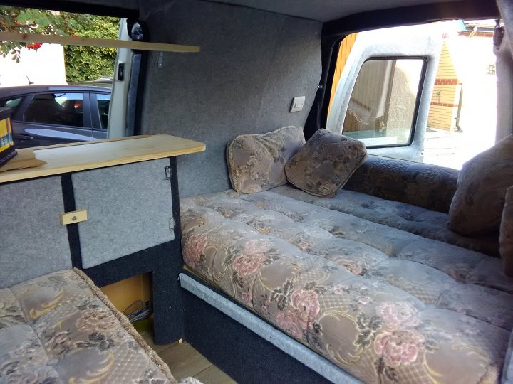 A bedroom with a bed and a table - The image shows the interior of a large van or motorhome, which has been customized with upholstery. The main seating area features a adjustable bench with a floral-patterned seat cover that is folded down. Next to this area, there is a raised area with a matching seat and a flat, surfboard-like surface that may serve as a desk or shelf. Above this surface, there is an open storage compartment. The vehicle is parked in a location with other vehicles visible, and there appears to be some construction or renovation activity happening in the background.