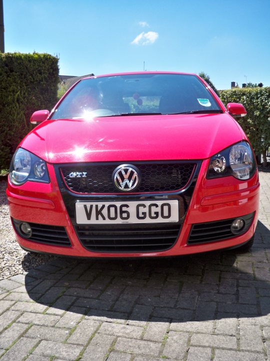 Pistonheads Polo Gti - The image is a photograph of a red Volkswagen car under a clear, blue sky. The car features a reflective grille and modern, angular headlight designs. It is parked on a brick pavement, casting a shadow on the ground. There is a watermark on the front grille with the logo of the insurance company "TuVan!" and a registration plate that reads "VK06 GGO." The image has a bright and sunny atmosphere, with the car being the central subject of the image.