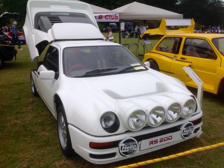 What is your daily?! - Page 9 - Supercar General - PistonHeads - The image showcases a vibrant scene at a car show or event. Dominating the left side of the image is a large white coupe equipped with a pair of aftermarket blue spotlights, giving it a distinctive and stylish look. The coupe is parked on grass with other vehicles in the background, suggesting a unique outdoor setting for the event. There aren't any people directly visible in this view, but there are some people in the background and beside the vehicles, indicating the presence of attendees. There are also some chairs and other items set up in the background, suggesting a relaxed and informal atmosphere.