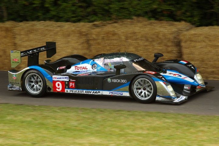 FOS good photography vantage points.. - Page 1 - Goodwood Events - PistonHeads - The image features a sleek, blue and black LMP2 racing car with the number 9, sponsored by Total and Peugeot. The car is prominently displayed on a grassy track. The setting suggests a racing event, as there is a hay bale barrier in the background. The number 9, along with its sponsors, is visible on the car's body. The background is filled with a lush green, creating a stark contrast with the blue and black color scheme of the racing car.