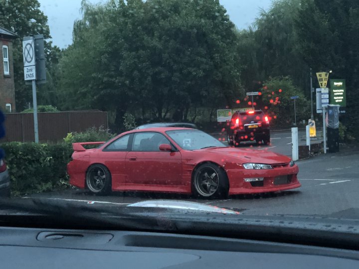 Pictures of decently Modified cars [Vol. 2] - Page 300 - General Gassing - PistonHeads - The image depicts a scene on a wet road at night or twilight. There's a red racing car parked on the side of the road, facing away from the viewer. In the background, there are trees, a pedestrian crossing marked by reflective strip, and a sign. Another car is seen driving away from the camera. The image captures the reflection of the red car on the car's interior window, indicating the photo was taken from inside another vehicle. All the cars appear to have their headlights on. The overall impression is of an urban or suburban environment.