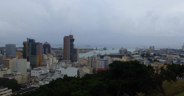 A large clock tower towering over a city - Pistonheads - The image captures a broad view of a cityscape under a grayish sky, suggesting an overcast or possibly cloudy day. Dominating the foreground are a variety of buildings, including a Coca-Cola Company building, which are densely packed. The city appears to be situated on the outskirts of a large body of water. In the distance, several boats can be spotted, hinting at an active harbor. The overall scene suggests an urban setting with a significant presence of commercial structures.