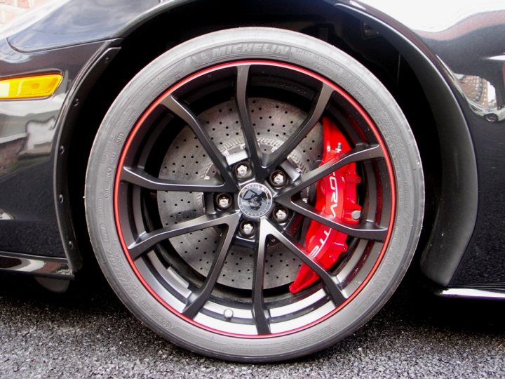 Corvette Z06 Centennial Edition for sale in the UK - Page 1 - Corvettes - PistonHeads - The image features a close-up view of a car's wheel. It has a shiny silver-colored center with a series of small holes that could suggest a drilled rotor. The outer rim of the wheel is black, with a wide area of red at the bottom, reminiscent of a Ferrari logo design. The spokes connecting the center to the outer rim are black with a complex, mechanical pattern. The wheel is resting on a gravel-like surface, providing a textured background to the scene.