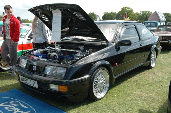 Fins and Chrome - Page 1 - North East - PistonHeads - In the image, a black Ford Granada sports coupe is the main focus. It has a striking blue engine intake system. The car is parked on a grass field during a motor show, as indicated by a blue Ford emblem next to the car. Two people can be seen, possibly bystanders enjoying the display. In the background, there are other vehicles parked, adding to the bustling atmosphere of the motor show. The car and its surroundings exhibit a sense of commitment to high-end, performance-oriented automotive experiences.