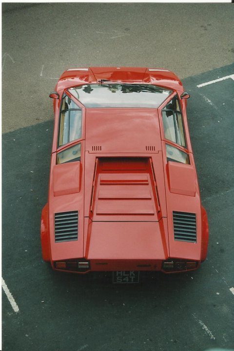 Countach  - Page 13 - Lamborghini Classics - PistonHeads - The image captures a striking red car, which dominates the scene with its vibrant color. This car is a convertible, characterized by its large rear window that offers a clear view into the vehicle. The model seen here is a Lamborghini Countach, a classic symbol of sports car luxury and performance. The photograph is taken from a high angle, providing a wide perspective of the car as it is positioned in a parking lot. The car is parked on a tarmac that extends to a black fork diagonally, leading the viewer's eye towards the horizon.