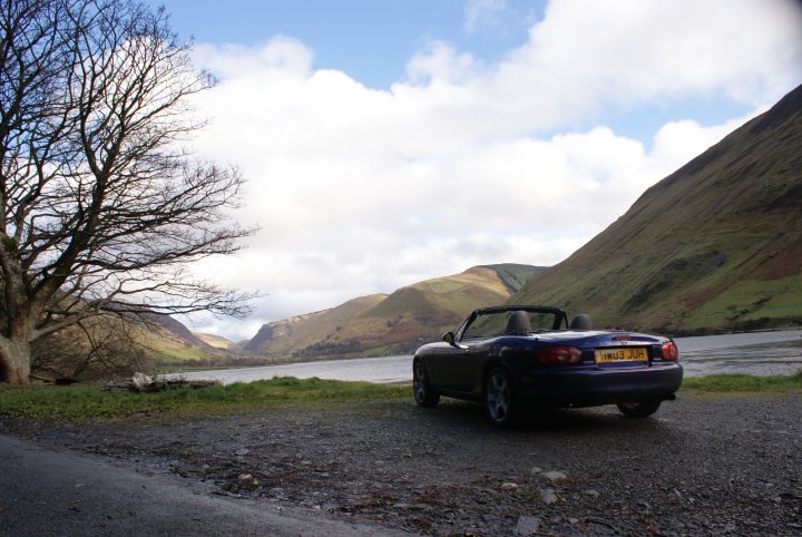 Roadster Roadtrip - Page 1 - Mazda MX5/Eunos/Miata - PistonHeads - The image shows a tranquil scene of a car parked near a small body of water. The car, which is blue, is the main subject in the image and is situated on the right side of the scene, facing the camera. The sky above the car is filled with light blue hues that are speckled with faint white clouds. The foreground features a tree with bare branches indicating it might be autumn or winter. The landscape is lush and green with multiple hills or mountains in the distance under the cloudy sky. The overall atmosphere of the image is serene and peaceful.