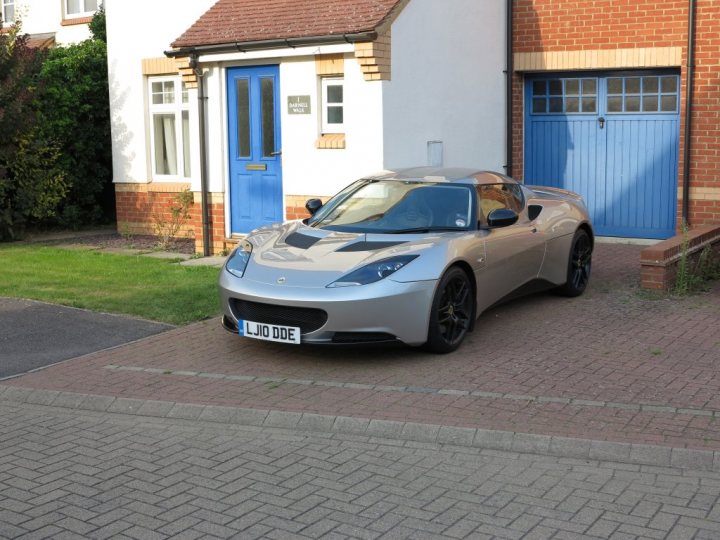 I've also bought one.... - Page 1 - Evora - PistonHeads - The image features a modern two-door sports car parked in the driveway of a two-story house. The car is gray and sleek, with a visible license plate reading "LIJ0DE." The house has white walls with blue doors and window frames. The garden area is neatly maintained, and there's a pathway lined with bricks leading up to the house. The overall atmosphere seems peaceful and residential.