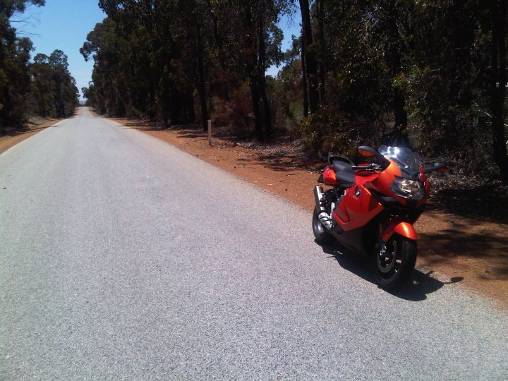 Phers Aussie Pistonheads - In the image, an orange and black motorcycle is parked on the side of a long, paved road. The road stretches into the distance and is flanked by a line of trees. The motorcycle's striking colors contrast with the natural hues of the trees, which display a variety of greens. The sky above is clear, suggesting a bright and sunny day. The motorcycle itself appears to be in good condition, with its bright contrast making it stand out against the more muted colors of the road and trees.