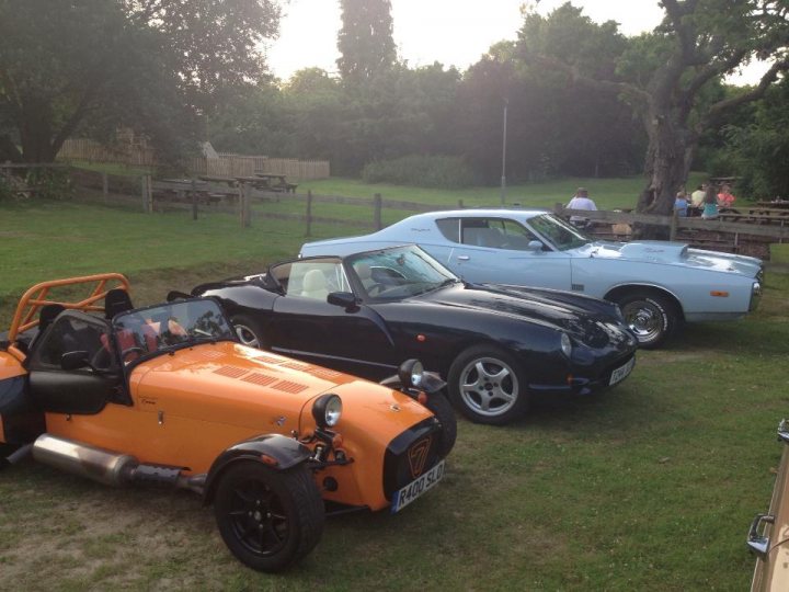 South Bucks Car meet Tuesday 13th August - Page 1 - Thames Valley & Surrey - PistonHeads - The image showcases a trio of vintage cars and a motorcycle parked on a grass surface, possibly in a rural setting due to the trees and benches in the background. The left side of the image features an orange car with a convertible top and emblazoned with various numbers, perhaps indicating racing or model designation. To the right sits a black vintage car, and in the center, there's a blue aerodynamic car that stands out with its sleek design. Behind these classic vehicles, a motorcycle adds to the diverse display of antique automobiles. The overall scene suggests a gathering or showcase of vintage automotive enthusiasts.