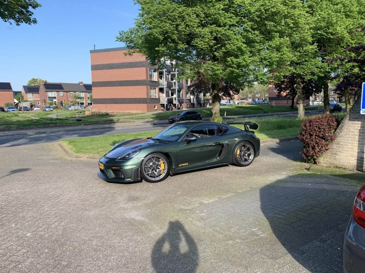 Pistonheads - The image shows a green sports car parked on the side of a street, with its hood up. It is positioned near a white parking sign, indicating that it is in a designated parking space. In the background, there is an urban setting with trees, buildings, and a blue sky visible above. There is also a reflection of the photographer in the car's shiny surface. The photo seems to be taken during the day under clear skies.