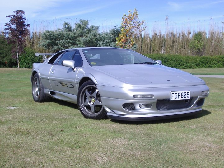 My old Esprit.. - Page 2 - Esprit - PistonHeads - The image features a silver Porsche 911 GT2 sports car, parked on a grassy field. The car is dominated by a large rear wing and is adorned with a yellow decal on the hood. The car's sleek design and tailored body hint at its high performance capabilities. The field it's parked on is vast and open, and beyond it, lush trees stretch towards a clear blue sky. The scene is serene, with the sun shining brightly, casting a soft glow on the car and highlighting the snow on its roof, indicating recent weather conditions.