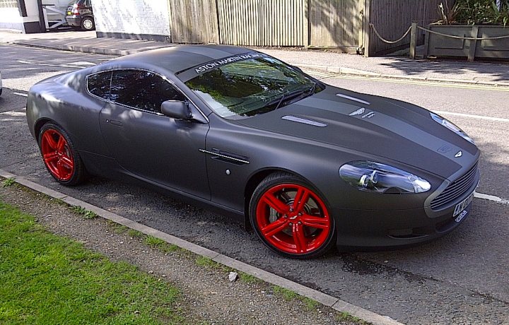 alloy wheel re-furb needed - Page 1 - Aston Martin - PistonHeads - The image shows a silver sports car parked on the side of a road, viewed from the rear passenger side. The vehicle has a sleek, streamlined design, featuring red alloy wheels and a complex body pattern with numerous vents and creases. The car is positioned next to a curb, which appears to be made of grass, indicating that this might be a residential or park-side location. There is a portion of a chain-link fence visible in the background. The lighting suggests it is a sunny day, with bright shadows cast on the car and the ground.