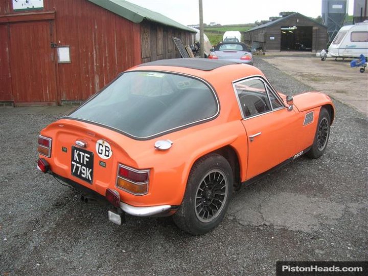 Early TVR Pictures - Page 65 - Classics - PistonHeads - The image depicts a vibrant orange classic car, identified by the "KTF 77K" license plate, parked in a gravel lot. The car is adorned with a rally style number plate. In the background, a dilapidated building adds a rustic charm to the scene, contrasting with the shiny, well-maintained appearance of the car. The contrast between the old, proud vehicle and the aged building evokes a sense of nostalgia and timeless beauty.