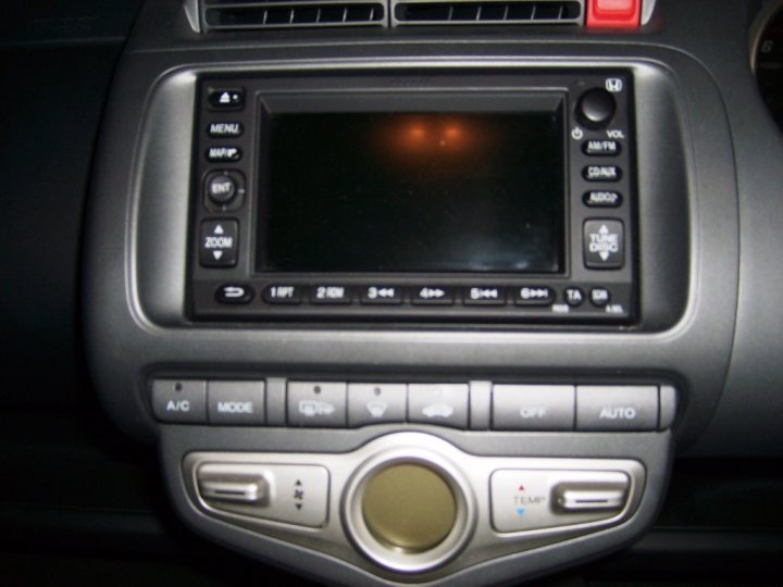 Honda Pistonheads Honest Theyre Jazz - The photo shows an interior view of a vehicle, focusing on a car stereo system. The stereo unit is a touchscreen type, centrally positioned on the car's dashboard. The screen is currently off, reflecting the static of a recently turned off device. Surrounding the screen are several buttons, some of which appear to have symbols on them, possibly for changing settings or navigating through menus. The stereo interface also has a clock and temperature display, providing information about the current time and thermal conditions inside the vehicle. The background, though not sharply in focus, suggests a steering wheel and part of the dashboard, reinforcing the vehicle's ambiance. The image has a shallow depth of field, with the lights reflected on the screen and the buttons slightly out of focus.