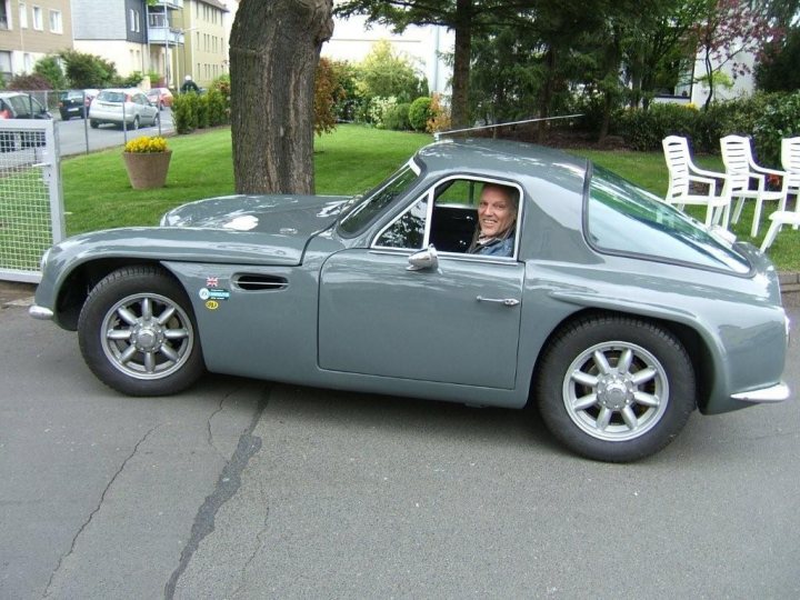 Early TVR Pictures - Page 11 - Classics - PistonHeads - The image showcases a man joyfully driving a gel blue Mercedes 320. He is seated in the driver's seat, smiling towards the camera. The car is parked on a street with parked cars in the background. The setting is bright, and the man is surrounded by nature, with a bench visible in the frame. The overall scene evokes a sense of pleasure and adventure.