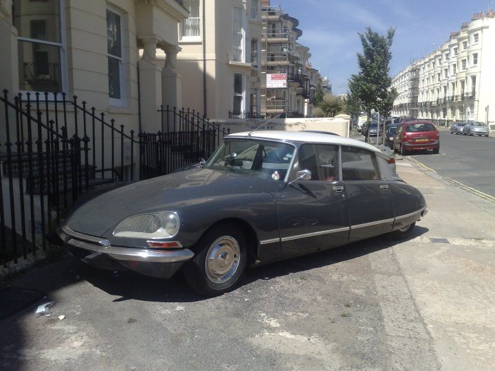 Pistonheads - This image shows a classic, vintage car parked near the curb on a city street. The car appears to be a two-door model with a prominent crest in the hood area. The vehicle is predominantly gray and has round headlights. It's positioned in front of a building with a black gate, and the street is lined with parked cars and residential buildings. There are no visible license plates or distinctive marks that provide further identification or location. The setting seems ordinary, typical of many urban areas with a sky that is clear, indicating a pleasant, sunny day.