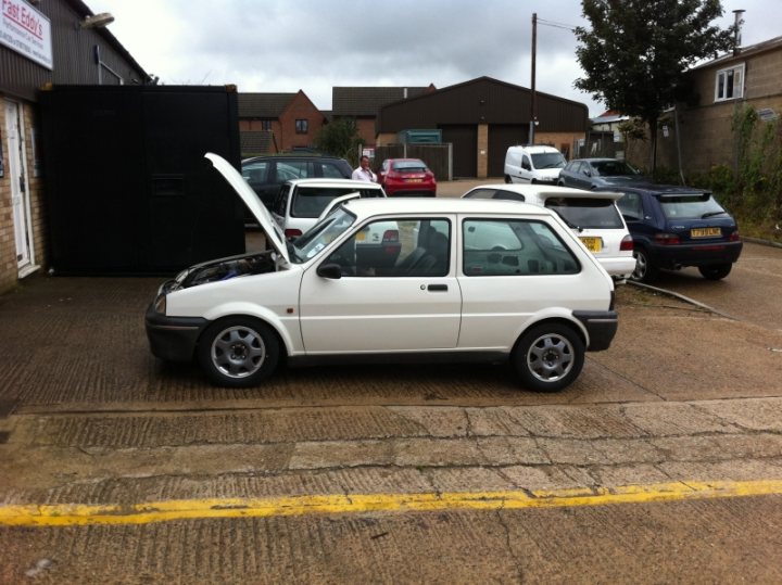 New Car Sneak Preview - Page 1 - East Anglia - PistonHeads - In the foreground of the image, there is a yellow line painted across a road or parking area. Behind this line, there is a white hatchback parked in a parking lot or on top of a road. The car has its hood open, revealing the engine. The lot appears to be a mix of commercial or industrial buildings. There are cars parked both in the foreground and the background, but the white hatchback with its hood up is the central focus of the image.