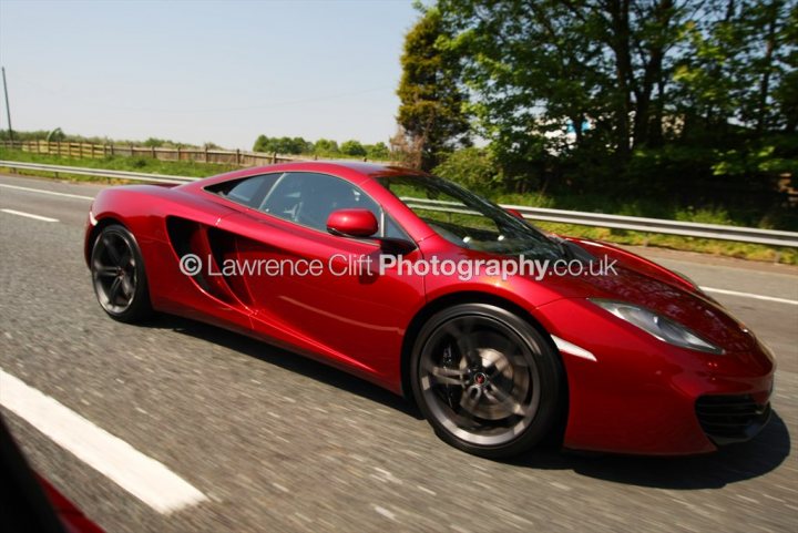 A red and white car parked in a parking lot - Pistonheads - The image shows a red sports car moving down a road. The car's sleek design and the shiny finish suggest it's a high-performance vehicle, possibly a sports car or a luxury convertible. The photo captures the dynamic feel of the car's motion, as evidenced by the blurred background and the silhouette of the photographer's hand holding the camera to the window.