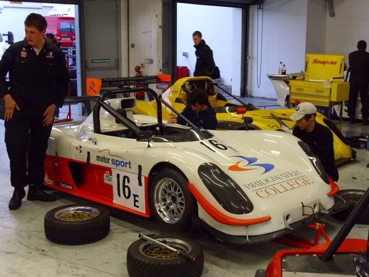 Association Day Picturs Stroke Pistonheads - This image showcases a bustling scene in a large garage or warehouse. There are two race cars parked next to each other. One is a bright yellow machine with a black seat. The other is a white car adorned with blue and orange accents and a blue and orange coat of arms symbol on the side.

Around these cars, there are two individuals. One is dressed in a black top and is standing near the white car, while the other is wearing a black hat and is standing next to the yellow car. They seem to be engaged in work or inspection of the cars.

In the background, there's a red truck, a blue water cooler, and a white table. The presence of these objects suggests that this is a professional setting, possibly a racing facility or a car repair workshop.