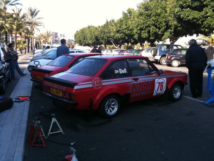 Rally Isla Pistonheads Mallorca Clasico Vii - The image showcases a street scene where two red cars with a racing theme, are parked next to each other. The parking lot is moderately populated with other cars and there are people standing around. The vehicles are adorned with bumper stickers that read "Bell", indicating they might be racing cars or used for racing purposes. The cars are parked on a paved road in broad daylight, with palm trees visible in the background, suggesting a tropical or subtropical location.